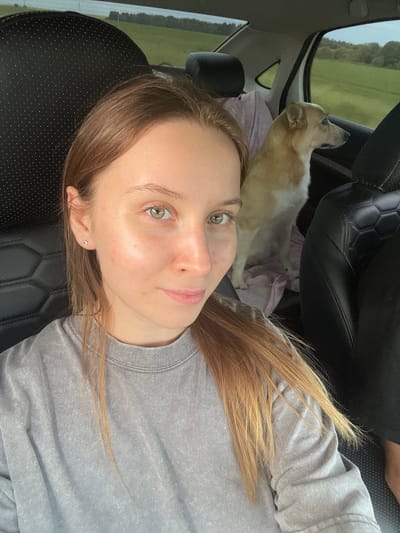 A young woman with blonde hair and green eyes is taking a selfie in the back seat of a car. She is wearing a gray sweatshirt. A light brown and white dog is sitting next to her, looking out the window. The car is on a road in a rural area, with green fields and trees visible outside. The interior of the car is dark leather. The image was taken in Povarenki, Russia.