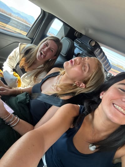 Three young women are seated in a vehicle. The woman in the foreground smiles with braided hair and a navy top. The middle woman, blonde with a seatbelt, purses her lips. The back woman sticks her tongue out, wearing a light jacket. The scene is bright, and the background shows a blurred landscape through the car window. Interior car details are visible.