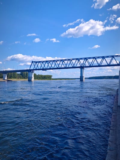 The image shows a wide, blue river with choppy water under a bright blue sky with scattered white clouds. A large blue truss bridge spans the river from left to right. The bridge is supported by concrete pillars. On the far bank, a line of green trees is visible. On the right side of the image, a concrete embankment or walkway runs along the edge of the water. The scene appears to be a sunny day, likely in Abalakovo, Russia, given the presence of the bridge which is a prominent landmark there. There are no people visible in the image.