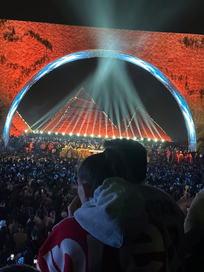 The image captures a nighttime scene at the Giza Plateau in Egypt, likely during a sound and light show or special event. In the background, a pyramid is illuminated with lights, creating a distinct triangular shape with horizontal lines. Beams of light project upwards from the pyramid.

A large, rounded arch or portal frame is positioned in front of the pyramid. The arch is lit with a bright, blue light, creating a halo effect. The immediate surroundings of the archway display a reddish-brown textured surface, possibly representing ancient stonework or structures.

The foreground shows a crowd of people, many of whom appear to be using mobile phones, presumably to capture photos or videos of the scene. The person wearing a jacket with a number on it is facing the pyramid along with the crowd of people. The general ambiance suggests a popular event or spectacle drawing a large audience.