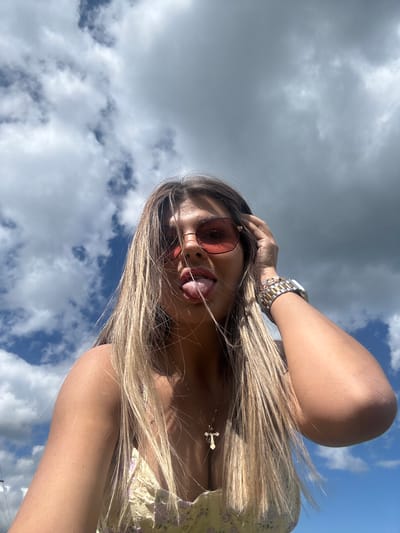 In a close-up, low-angle selfie, a young woman with long, blonde hair is pictured outdoors against a backdrop of a dynamic sky with both blue patches and grey clouds. She is wearing rose-tinted sunglasses and a yellow floral top, with a cross pendant necklace visible. Her right hand is raised, touching her hair, and her tongue is sticking out. A silver and gold watch adorns her left wrist. The image does not provide any specific details to identify a location in the city of Пастухово, Russia.
