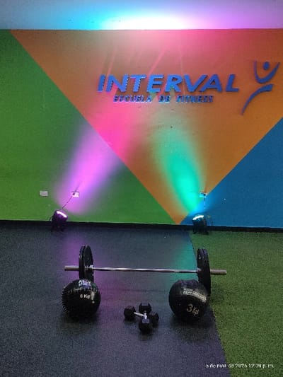 The image depicts the interior of "INTERVAL ESCUELA DE FITNESS" in Quilmes, Argentina. The background wall is divided into large geometric sections painted green on the left, orange in the center, and blue on the right. The business name, "INTERVAL ESCUELA DE FITNESS," is displayed in raised silver-blue letters on the orange section of the wall, with a stylized blue human figure logo to its right. The wall is illuminated by colored spotlights, casting pink/purple light from the left and green/teal light from the right. Two black stage lights are visible on the floor against the wall, one under each colored light source. The floor consists of two distinct coverings: dark grey speckled rubber matting on the left side and green artificial turf-like matting on the right side. In the foreground, a silver barbell with black weight plates on each end is positioned horizontally on the floor. A black slam ball marked "ALLY SPORTS 5 Kg" is located to the left of the barbell, and another black slam ball marked "HIGH PERFORMANCE 3 Kg" is to its right. Two black hexagon-ended dumbbells are situated between the barbell and the slam balls, resting on the dark grey matting. No individuals are present in the scene. A timestamp in the lower right corner indicates "5 de mar. de 2024 12:39 p. m."