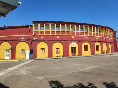 The image shows the exterior of a bullring under a clear, bright blue sky, suggesting a daytime scene with good weather. The building is predominantly painted in bold red and yellow, with a tiled roof. The lower level features a series of arched openings, some of which contain doors or windows, while others are open. The upper level has a covered walkway with a row of rectangular openings framed by yellow pillars and lintels. Some of the windows on the lower level have red bars. The ground in front of the bullring is paved with asphalt, and a white painted line runs across it. The overall impression is of a functional and perhaps historic building, possibly in a Spanish town given the location context. There are no people visible, and the scene appears quiet. There is some subtle greenery growing along the base of the bullring. There are a few utility boxes and lights mounted on the exterior walls. On one of the yellow arches, there appears to be a plaque or sign with some writing on it, but it is not legible. Another sign, a circular red and white sign, is mounted near a white door.