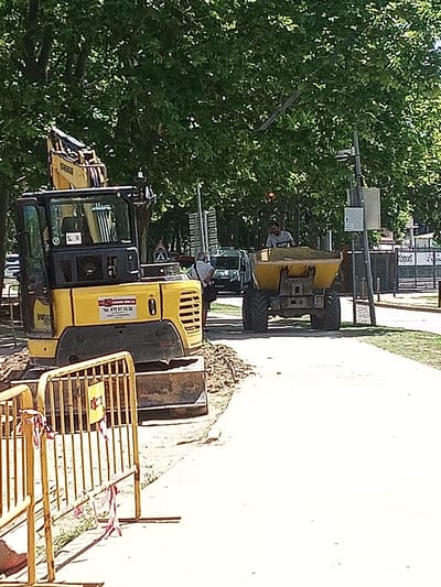 A yellow excavator is parked on a dirt path beside a paved walkway. The excavator has a black cab with windows and a yellow arm. A sign on the side of the excavator has the text "Tel. 972 57 15 35". In the background, a yellow dumper truck is being operated by a person. The dumper truck is filled with dirt or debris. The scene is outdoors, with trees providing shade. The time of day appears to be daytime, and the weather seems sunny. A yellow metal barrier with red and white tape is in the foreground, indicating a construction or restricted area. There are other vehicles and structures in the background, including what appears to be a sports facility. The overall impression is of a construction site in a park or public area.