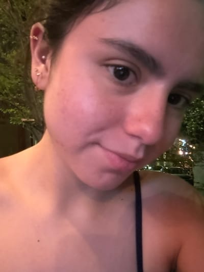 The image displays a close-up, self-portrait-style photograph of a young female subject, captured at night in an outdoor setting. The subject, appearing to be in her late teens or early twenties, features fair skin with some redness on her cheeks, dark eyes, and pronounced dark eyebrows. Her dark hair is pulled back and visible at the top and left side of the frame. She is looking directly at the camera with a subtle, closed-mouth smile or smirk, and her head is slightly tilted to her right.

Her visible left ear is adorned with multiple gold-colored piercings: a small hoop or cuff on the helix, a pearl-like stud in the conch, and two small hoops in the lobe. She is wearing a dark-colored (likely black or dark blue) spaghetti-strap top, exposing her left shoulder and clavicle. A visible tan line is present on her left shoulder.

The background is heavily blurred, indicating a shallow depth of field. On the left side, out-of-focus green foliage suggests trees or bushes. On the right, various indistinct artificial light sources are visible, consistent with streetlights, buildings, or vehicle lights, illuminating the scene with a warm glow. The environment is indicative of an urban or suburban area after dark. The precise location within the city of Santiago, Mexico, cannot be determined from the visual information provided in the image.