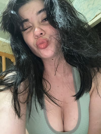 A woman with dark hair and a light-colored tank top is captured in a close-up selfie. She is winking and puckering her lips, with her hair framing her face. The background appears to be indoors, with hints of a wooden cabinet on the left and a patterned wall on the right. There is no discernible information within the image to indicate a specific location in Gavrilov Posad, Russia.
