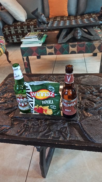 The image displays an indoor scene, primarily focusing on a collection of snack items and beverages resting on a heavily carved, dark wooden surface. The image appears to be rotated 90 degrees clockwise from its natural orientation.
When viewed upright, in the foreground, there are two bottles and a bag of potato chips. A brown glass bottle stands vertically at the top, featuring a red and white label that clearly reads "MKUNGA" and "MIST." A white 'V' symbol is also visible on its neck. Below this, a green bag of "Winnaz" brand potato chips lies flat. The bag is green with a prominent red and yellow logo, and it explicitly states "PAPRIKA" and "Potato Chips," accompanied by graphics of red paprika peppers and potato chips. Beneath the chip bag, a green glass bottle lies horizontally. Its label is white and red, and parts of the word "SULTZIG" are legible, along with other obscured text.
These items are arranged on an ornate, dark brown wooden surface, possibly a side table, tray, or a decorative panel. This surface features intricate, raised carvings with organic or abstract patterns.
The background to the left of the main focus reveals a relaxed living space. A grey sofa or couch is visible, adorned with various cushions, including a vibrant orange throw pillow and a lighter-colored, possibly white, pillow. Next to the sofa, a tall, vertical piece of furniture or a decorative screen stands, covered in a geometric patterned fabric or wood, showcasing colors like dark brown, red, green, and black, suggesting an ethnic or tribal design. Behind this, a dark, abstractly carved wooden object, possibly a mask or sculpture, leans against a wall. Below it, a small stack of books is neatly arranged. The wall in the background is a light, neutral color, possibly beige or off-white. To the far right, another section of the carved wooden structure is visible, forming a decorative bracket or support.
The overall impression is that of a comfortable living area, possibly a lounge, where snacks and drinks are casually laid out, suggesting a moment of relaxation.