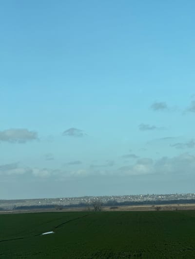 The image displays an expansive outdoor landscape under a predominantly light blue sky with scattered light gray and white clouds. The foreground consists of a dark green agricultural field, likely cultivated, featuring a small, irregular patch of white material, consistent with snow or ice, in the lower-left quadrant. Faint linear indentations are visible across the field. In the midground, a horizontal band of bare deciduous trees and brush, appearing light brown and gray, separates the field from the distant horizon. On the horizon, a low-density settlement, consistent with a town or village, is visible as a cluster of buildings, some with discernible pointed roofs, stretching across the landscape. No people, subjects, or active interactions are depicted. This scene is located in the city of Zarevo, Russia.
