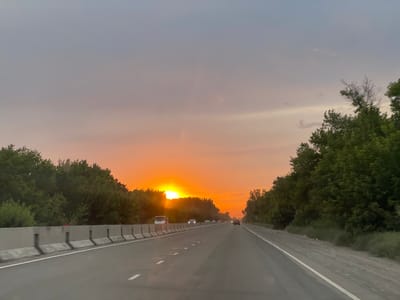 The image depicts a sunset over a highway, likely in Kyrgyzstan, given the location context. The sun is a brilliant orange orb, low on the horizon, casting a warm glow across the sky and the landscape. The sky itself is a gradient of colors, from a soft pink and orange near the sun to a muted grey-blue higher up, with wispy clouds adding texture.
The highway stretches into the distance, with cars and trucks visible, suggesting ongoing traffic. A concrete barrier with regularly spaced posts separates the lanes of traffic from the roadside vegetation. Dense green trees line both sides of the highway, forming a natural border. The time of day is clearly sunset, indicated by the low angle of the sun and the warm, diffused light. The weather appears clear, with the exception of the clouds.
There is no visible text in the image. The overall mood is one of calm and transition, typical of a sunset scene on a journey. The perspective is from inside a vehicle, looking forward along the road.