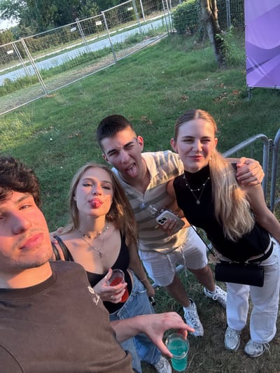 In this low-angle, slightly tilted selfie-style photograph, four young adults are gathered on a grassy area, likely at an outdoor event or festival. The sun casts a warm glow, suggesting it's late afternoon or early evening. The individuals are posed close together, with three of them looking directly at the camera.
The two individuals in the foreground, a young man and a young woman, are closest to the lens. The young man, with dark, curly hair, is in the bottom right corner, his mouth open slightly as if speaking or laughing. The young woman, with blonde hair, is positioned slightly behind and to the left of the man, sticking her tongue out playfully. She's wearing a black top and a silver necklace.
Behind them, two more individuals are visible. A young man with short, dark hair is on the left, also sticking his tongue out with a playful wink. He's wearing a striped shirt. To his right, a young woman with long blonde hair is smiling at the camera, wearing a black top and a pearl necklace. She holds a smartphone in her hand. Another person, whose face is not fully visible, is partially obscured by the others in the bottom right, holding a bright blue drink in a plastic cup.
The background shows a grassy field with some trees and a chain-link fence, indicating an outdoor setting. A large purple banner with a white design is partially visible in the top left corner. The overall mood is casual and joyful, with the subjects appearing relaxed and engaged in a social gathering. The image is oriented such that "up" is towards the left side of the frame.