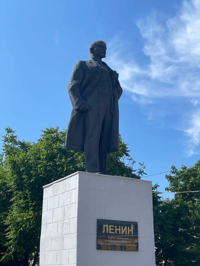 A full shot captures a dark, bronze statue of Vladimir Lenin standing on a tall, white, rectangular pedestal in Zavolzhye, Russia. The statue faces slightly to the left, with Lenin's right hand tucked into his coat pocket and his left hand holding the lapel of his jacket. He is dressed in a suit and overcoat. The pedestal has a plaque on its front with Cyrillic lettering. The background consists of a bright blue sky with wispy white clouds and lush green trees on either side of the statue. The lighting suggests it is daytime, likely a sunny day.