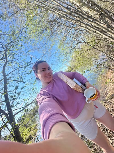 A young woman stands outdoors near a body of water, possibly a stream or river, surrounded by trees. The picture is taken from a low angle, looking up at the woman and the sky.
The woman is wearing a purple sweater and white shorts, with what appears to be a compression bandage on her thighs. She is holding a mug in one hand and a book in the other. She has a slight smile on her face.
The environment is a natural setting with trees and a water feature. The trees have new leaves budding, suggesting it is spring. The sky is blue and clear, with branches and leaves visible against it.
Based on the location provided, Черёмушки, Russia, this could be a park or wooded area within the city or on its outskirts.