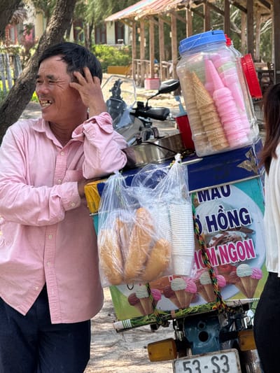 In Thuận Thành, Vietnam, a man in a pink collared shirt and dark pants stands next to a motorcycle-mounted ice cream cart. He is smiling and appears to be scratching his head. The cart is laden with goods, including a large plastic container filled with ice cream cones and what appear to be pink pastries or candies, as well as a plastic bag containing several loaves of bread. The side of the cart features colorful graphics advertising ice cream, with text in Vietnamese. The background shows a sandy ground, trees, and the structures of a roadside establishment.
