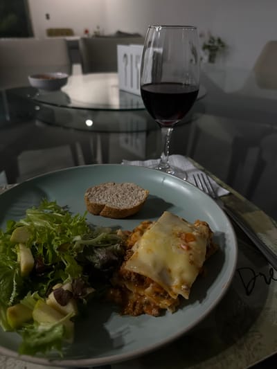 A plate of lasagna sits alongside a slice of bread and mixed green salad with sliced apples. A glass of red wine is placed next to the plate on a glass table. A fork and knife rest on a napkin above the plate. The background is blurry and shows chairs and other dining room elements.