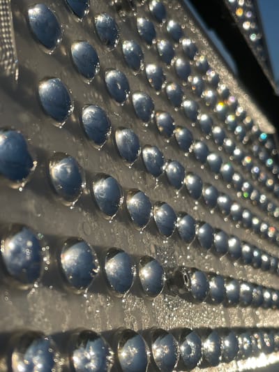 The image is a close-up, angled shot of a textured surface with rows of reflective, oval-shaped indentations. These indentations appear to be filled with a liquid, possibly water or dew, and they are reflecting the light from an unseen source, creating bright highlights and small, scattered rainbow refractions. The surface itself seems to be a dark, matte material, possibly metal or plastic, with a slightly grainy texture visible between the indentations. The lighting suggests it is daytime, with the sun low in the sky, casting long shadows and creating a sparkling effect on the wet surface. There are no people or specific objects identifiable in the frame, only the repeating pattern of the textured surface and its reflective qualities. The environment is abstract due to the close-up perspective, but the reflections hint at an outdoor setting. The image does not contain any specific visual cues to pinpoint its location within the city of Торковичи, Russia, or any other specific geographical location.