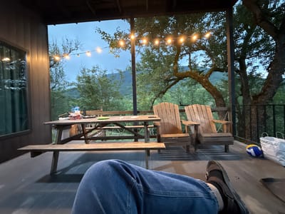 The image depicts a porch area at dusk, overlooking a natural landscape with trees and distant hills. A person's legs, clad in blue jeans and dark sandals, are positioned in the foreground, indicating they are seated or reclining on the porch. In the midground, a wooden picnic table with benches is set up. On the table are several items, including bottles of water, a red container, and what appears to be a green plate. Two Adirondack-style wooden chairs are placed to the right of the picnic table. Further to the right, a volleyball and a white shopping bag are visible on the porch floor. The porch is illuminated by a string of fairy lights overhead, casting a warm glow. The overall scene suggests a relaxed, outdoor setting, possibly a vacation rental or a residential property. The image does not contain any visual information that would allow for the identification of the specific location within the city of Sis, Azerbaijan.