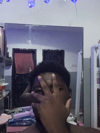 The image depicts an indoor scene in Gbongan, Nigeria, captured as a mirror selfie. A dark-skinned individual with short, dark, curly hair partially obscures their face with their right hand, revealing only their left eye. The person's bare shoulder is visible in the lower right foreground.
Reflected in the mirror, the background shows a room with light-colored walls. Blue decorative lights are strung across the upper portion of the reflected wall, casting a blue hue and creating small, scattered reflections resembling glitter on the mirror surface. Two dark, rectangular openings or doorways are visible in the reflection, with a reddish-brown towel draped over the left opening. To the far reflected left, a dark cylindrical object rests on a shelf. To the far reflected right, a white shelving unit is partially visible, containing various items including a bottle with text "Spra" on its label.
The surface directly in front of the mirror, below the individual's reflection, displays several objects: a clear container with white contents, a blue and white packaged item (possibly wipes), a light pink object (resembling a small fan or brush), and a light pink lid in the bottom left.