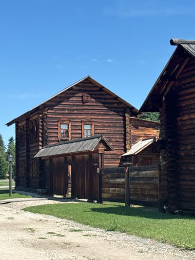 The image depicts a rustic, traditional wooden building complex under a clear blue sky in Shushenskoye, Russia. The primary structure is a large, two-story log cabin with a steeply pitched roof, characterized by its horizontal logs and visible timber construction. There are several windows on both levels. A wooden entryway with a roofed porch and double doors is positioned in front of the main building. To the right, another log structure, partially visible and at an angle, appears to be part of the same compound, with a connecting wooden fence. The surrounding area features a gravel path winding through a grassy lawn, with evergreen trees visible in the background. The overall scene evokes a sense of historical preservation and rural architecture.