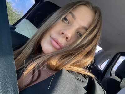 A close-up, selfie-style shot shows a woman from the chest up, seated in the passenger seat of a car. She has long, light brown hair that partially covers her face. Her eyes are a greenish-brown, and she is wearing light makeup. A silver necklace with a crescent moon pendant is visible. She is wearing a black jacket. The background shows the car's interior, including the headrest, seatbelt, and a quilted white pillow on the seat. Outside the car window, trees and a blue sky with clouds are visible. The location is identified as the city of Kuznechikha, Russia.