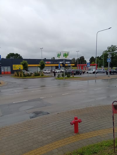 The image depicts an overcast day in Kuršėnai, Lithuania, specifically in the vicinity of an IKI supermarket. In the foreground, a red fire hydrant stands on a paved walkway with a textured yellow tactile paving strip. Beyond the walkway is a wet asphalt road with white lane markings and some patches of standing water, indicating recent rain. Across the road, the IKI supermarket building is visible, with its distinctive green logo. Several cars are parked in the supermarket's lot. Pedestrian crossing signs with figures walking are posted on poles, and a traffic light is also present. A few individuals are seen walking in the parking lot area. The overall environment appears to be a commercial district with a functional, everyday atmosphere.