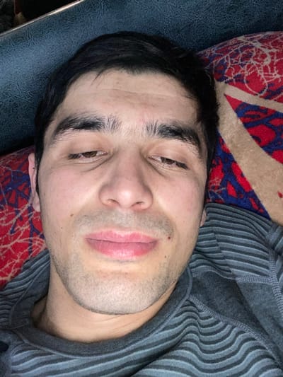 A fair-skinned man with dark hair and eyebrows lies on a patterned surface, possibly a cushion or blanket. His gaze is slightly downward, and his lips are closed. He has stubble on his face. He wears a gray striped top. The background consists of patterned fabric with red, blue, and beige colors. The overall impression is one of relaxation or repose.