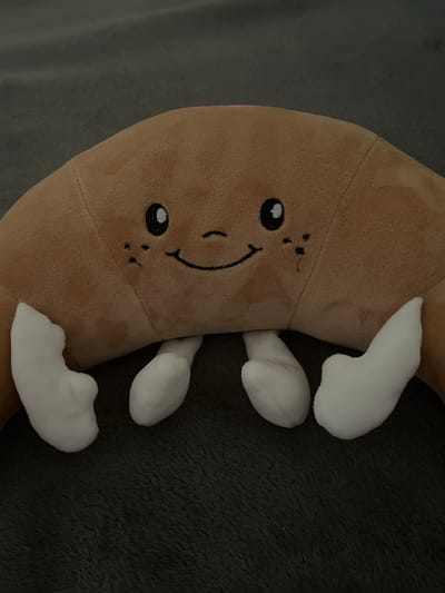 A plush croissant toy is lying on a dark gray, textured surface. The toy is a crescent shape, made of light brown fabric. It has a smiling face with two black eyes, small black freckles on each cheek, and a black stitched mouth. Four white, stubby feet are attached to the underside of the toy, protruding outwards. The surface it rests on appears to be a blanket or rug. The image is an indoor close-up shot, and no specific location in Olomouc, Czechia is identifiable.