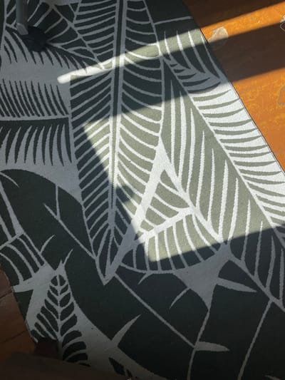 An overhead shot displays a dark green and white patterned rug with a leaf motif. The rug is partially covered by sunlight casting distinct shadows. To the left of the rug, a dark grey metal leg of a piece of furniture with a black caster is visible, partially obscured by shadow. The rug lies on a brown wooden floor. The image was taken in Kopeysk, Russia.