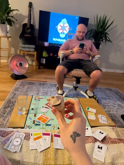 The image captures a casual indoor scene. A person sits in an exercise chair in front of a television displaying the word "SABAB" along with an abstract visual, and has a cell phone in their hands. A guitar leans against the wall next to the television, and there's a small space heater on the floor. In the foreground, another person is seated at a Monopoly board, with property cards and money visible. A stone sculpture of a face is being held over the board, seemingly acting as a playing piece. The scene is lit naturally, and a houseplant is situated in the background, suggesting a home environment. The image does not provide enough information to place the image within the specific city of Morton Grove, United States of America.