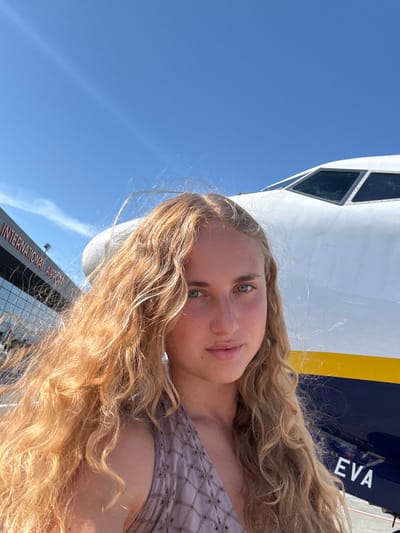 A young Caucasian woman with long, curly blonde hair and blue-green eyes is taking a selfie outdoors. She is wearing a light purple sleeveless top with a patterned texture. Behind her, a large white airplane with a dark blue fuselage and a yellow stripe is partially visible. The plane's cockpit windows are prominent. To the left, the exterior of a modern building with glass windows is visible, featuring red lettering that reads "INTERNATIONAL AIRPORT". The sky above is a clear, bright blue. The scene appears to be at an airport.