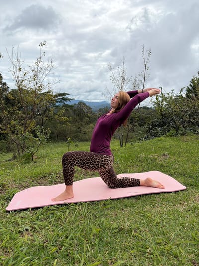 A woman with auburn hair is performing a yoga pose on a pink mat outdoors. She is wearing a plum-colored long-sleeved top and leopard-print leggings. Her right knee is on the mat, and her left leg is bent at a 90-degree angle with her foot flat on the mat. Her arms are raised above her head, with her hands clasped. She is facing to the left of the frame. The background shows a lush green hillside with trees and mountains in the distance under a cloudy sky. The location appears to be a natural setting, possibly in or near Ban Khun Wang, Thailand, given the vegetation and mountainous landscape.