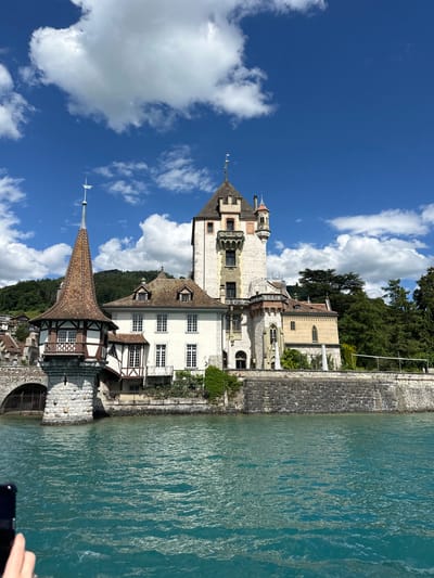 This is a vertical, eye-level photograph taken on a sunny day with a bright blue sky dotted with white, fluffy clouds. The scene depicts a picturesque view of Oberhofen Castle on the shores of Lake Thun in Switzerland.
The castle itself is a prominent subject, with its striking white facade and distinctive architecture. It features several towers, including a prominent square tower with a pitched roof and a spire topped with a flag. The castle boasts intricate details, such as decorative balconies with crenellations, arched windows, and gabled roofs. Some sections of the castle show a timber-framed style with white walls and dark wooden beams.
Adjacent to the castle, a section of stone wall lines the edge of the property, leading down to the tranquil turquoise waters of Lake Thun. The water is calm, with gentle ripples reflecting the sunlight, creating a shimmering effect. Lush green trees and dense foliage are visible behind the castle, suggesting a verdant landscape.
The time of day appears to be daytime, given the bright sunlight and clear sky. The weather is pleasant and fair, ideal for outdoor viewing. The overall mood is serene and idyllic, capturing the beauty of a historic Swiss castle set against a stunning natural backdrop. No people are clearly visible in the frame, and there is no visible text.