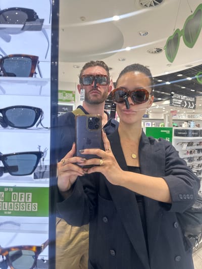 Two individuals, a male and a female, are taking a mirror selfie in what appears to be an opticians or sunglasses store. They are both wearing sunglasses in a humorous way, with the male's sunglasses positioned on his nose and the female's sunglasses also on her nose. The male is wearing a dark top with a khaki strap over his shoulder, while the female is wearing a dark pinstripe blazer over a dark top, accessorized with a gold necklace and rings. They are both holding a mobile phone to take the photo. In the background, there are displays of sunglasses and promotional signage, including one that reads "UP TO % OFF GLASSES". The environment suggests a retail setting, possibly within a shopping mall or airport, given the lighting and ceiling structure. There are no specific landmarks that would identify the location as Uttlesford, United Kingdom.