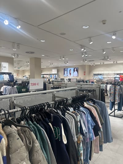 The image captures a well-lit interior of a retail clothing store, likely a fast-fashion chain, given the presentation of merchandise on racks. The store is located in the Polígono Industrial De Quart De Poblet, Spain. The scene is dominated by rows of clothing displayed on metal racks. In the foreground, a rack of casual wear, including puffer jackets and t-shirts, is visible. A prominent white sign displaying "7,99 €" is positioned in the mid-ground, indicating a sale price. Behind this, more clothing racks extend into the background, featuring a variety of garments like shirts, dresses, and jackets. Overhead, a modern ceiling with numerous track lights and spotlights illuminates the space. A large digital screen displaying fashion advertisements is mounted on a far wall. The flooring appears to be a polished concrete or tile. No individuals are discernible in the image. The overall impression is one of an organized and brightly lit retail environment stocked with apparel.