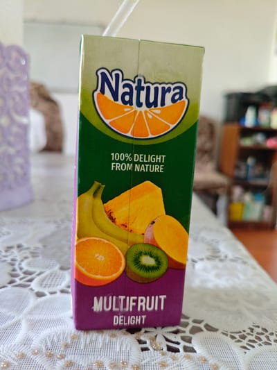 A close-up, eye-level, medium shot captures a purple and green juice box from the brand "Natura" standing upright on a white, lace-patterned tablecloth. The juice box features illustrations of various fruits, including an orange slice, bananas, pineapple, mango, and kiwi. Text on the box reads "Natura," "100% DELIGHT FROM NATURE," and "MULTIFRUIT DELIGHT." A white straw protrudes from the top of the juice box. The background is softly blurred, showing a wooden shelf with various items, and a portion of a wall. The overall scene suggests a domestic setting, possibly in a home in Sumgayit, Azerbaijan. There are no people visible in the image.