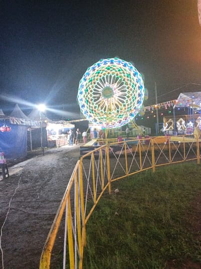 An outdoor night scene features a vibrant Ferris wheel with colorful lights. Tents and food vendors line a dirt path on the left. A yellow and white fence runs along the right, separating the path from a grassy area. People gather near the carnival attractions, illuminated by bright lights that pierce the darkness.