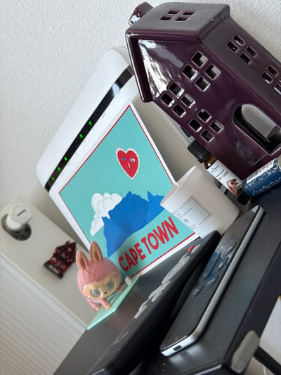 The image shows a collection of objects seemingly arranged on a flat surface, possibly a shelf or table. On the left is a purple ceramic house-shaped object with square cutouts for windows. Next to it, there's a white Wi-Fi router with several illuminated green indicator lights. Atop the router rests a card with the words "CAPE TOWN" in large red letters, along with a graphic of a blue mountain and a heart-shaped image with tears. A small white candle or container with Japanese writing is also present. Further to the right, there are what appear to be stacked electronic devices, possibly including a laptop and a remote control. Also present is a small, pink, anime-style figurine, and an aromatherapy oil bottle. A small decorative house and a dice are also visible on the surface. The location is not identifiable as 's-Hertogenbosch, Netherlands from the available details.