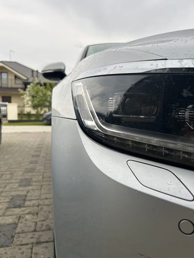 This is a close-up, angled shot of the front passenger-side headlight and bumper area of a silver car. The car appears to be parked on a paved driveway. Water droplets are visible on the car's surface, suggesting it might be raining or has recently rained, and the sky is overcast.
The headlight is a modern LED design with a prominent daytime running light strip. To the right of the headlight, there is a small, diamond-shaped cover, likely for a sensor or tow hook, and a circular sensor, possibly a parking sensor, is also visible on the bumper.
In the background, a residential house with a dark roof and windows is partially visible, along with some greenery from trees or bushes. The ground is paved with rectangular stones, some of which also show water droplets. The overall scene suggests an ordinary day, perhaps in the morning or afternoon given the lighting. No people are visible in the frame. There is some faint text visible on the headlight assembly, possibly a manufacturer's logo or model number, but it is too indistinct to read. The image is cropped tightly on the car's front, emphasizing its design and features. The location is identified as Haaslava, Estonia.
