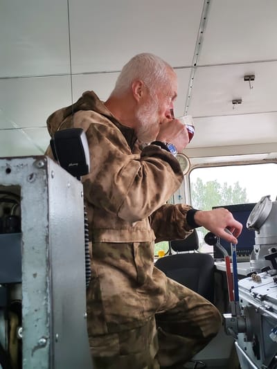 A bearded, white-haired man wearing a camouflage jumpsuit is seated inside a vessel's control room, holding a glass of red liquid to his lips. He is looking out a window, his left hand resting on a control lever with red and blue markings. To his left is a piece of machinery, and to his right are various control panels, a computer monitor, and a navigational instrument. The interior of the control room features a white ceiling with visible paneling and lighting fixtures. Outside the window, trees are visible under an overcast sky. The scene appears to be within the city of Vytegra, Russia, likely on a waterway such as the Onega Canal or Lake Onega, given the maritime setting.