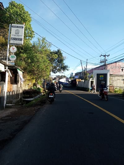 The image presents a daytime street scene in the city of Khouathineung, Laos, under a clear blue sky. A dark asphalt road, marked with a single yellow line, extends into the background, with a slight curve to the left.
On the left side of the road, a vertical sign post holds a prominent white rectangular sign with "FOOD COURT" at the top. Below this, the text "DISUDUT SEKOLAH" (Indonesian/Malay for "At the Corner of the School") is visible, followed by additional smaller text. Below the main sign are three circular signs featuring distinct logos: a red rooster, a green plant or flower, and three green stylized trees. Dense green trees and foliage line the left side of the road, partially obscuring a rustic-looking building structure.
On the right side of the road, a rectangular monument or sign structure with a shield-shaped crest (displaying yellow, green, and blue sections) is visible. Behind this, a light-colored building with the partial word "SIM" on its facade is present. A large utility pole with multiple transformers and numerous overhead power lines dominates the upper right. A group of three to four individuals stands near the monument.
Several motorcycles are active on the road. In the mid-ground, a person wearing a dark jacket and helmet rides a dark scooter away from the camera, displaying a license plate. Another dark motorcycle follows further ahead. On the right, a person in a dark helmet and light blue top rides a red motorcycle towards the camera. Additional motorcycles are visible in the distance. The scene is brightly illuminated by natural sunlight.