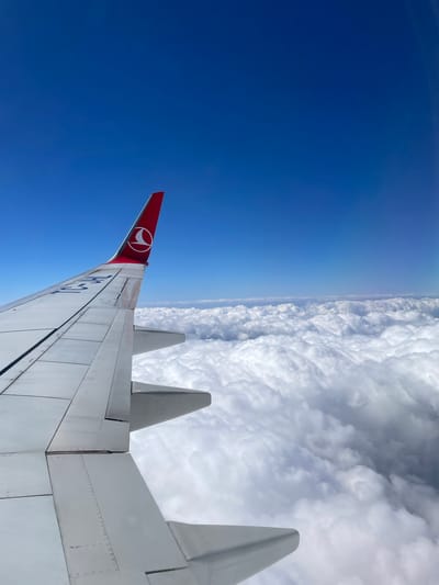 The image is taken from inside an aircraft, likely during flight. The view shows the wing of the plane, with a red tail fin bearing the logo of Turkish Airlines (THY). Below the wing on the left is a clear, deep blue sky. On the right, dense white clouds fill the frame. No people are visible. The aircraft's tail number is visible on the wing. The environment is the upper atmosphere. The city of Süleymanpaşa, Turkey, is not discernable.