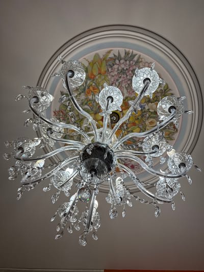 A crystal chandelier hangs from a painted ceiling in a room, possibly in Ozeretskoe, Russia. The chandelier is ornate, with multiple arms extending outwards, each adorned with crystal droplets. The ceiling features a circular mural with a floral and fruit motif, bordered by decorative trim. The lighting in the room is dim, allowing the chandelier to be the main focal point. The image is taken from a low angle, looking up at the chandelier and ceiling. There are no people or animals visible in the image.