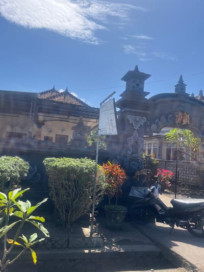The image captures a sunny outdoor scene in Jembrana, Indonesia, featuring traditional Balinese architecture. In the foreground, lush green bushes flank a dark-tiled pathway. Beyond the bushes, a weathered stone archway, intricately carved with cultural motifs and topped with decorative spires, dominates the mid-ground. A signpost with writing in Indonesian is attached to the left side of the archway. A motorcycle is parked to the right of the archway, partially obscured by colorful plants and a statue draped in a checkered cloth. In the background, a building with a tiled roof and decorative windows is visible. The sky above is a bright blue, with scattered white clouds and a few thin wisps of cirrus clouds.
