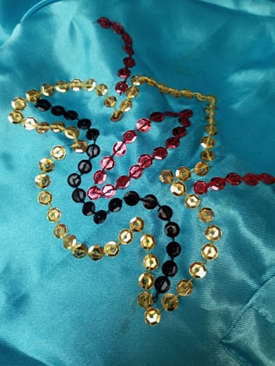 On a turquoise fabric background, a design is formed from strings of sequins in three colors. Gold, black, and red sequins are strung together in parallel lines, creating a curved and angular decorative pattern. The sequins catch the light, adding shine to the fabric. Slight discoloration is present on the material.