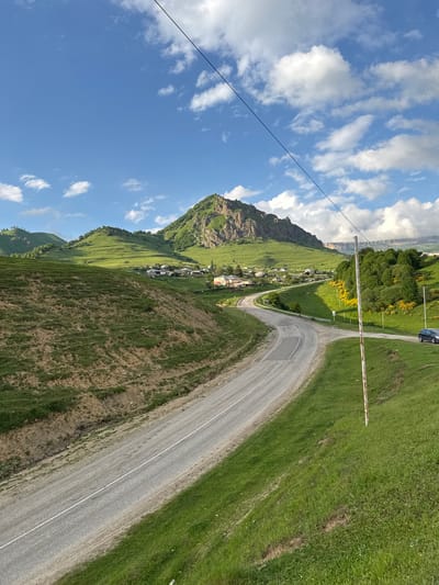 This image captures a scenic view of a winding road traversing a lush, green mountainside in Nizhnyaya Mara, Russia. The scene is bathed in the bright sunlight of what appears to be a clear day, with scattered white clouds dotting the vibrant blue sky. A power line runs diagonally across the upper left portion of the frame, adding a man-made element to the natural landscape.
The road, paved and marked with a white line, curves gracefully around the mountain. On the right side of the road, a verdant, grassy slope dominates the foreground, with patches of dry earth and some erosion visible. A red and white barrier pole is positioned across the road, suggesting a checkpoint or an entrance.
Further up the mountainside, nestled amongst the green slopes, a small village or settlement can be seen. Houses with red roofs are clustered together, giving a sense of community within the vast natural setting. The mountain itself rises dramatically behind the village, its rocky facade partially covered in greenery.
To the left of the road and mountain, the sky takes center stage, with fluffy white clouds creating patterns against the deep blue expanse. A black car is partially visible in the upper right corner, driving on the road. The overall impression is one of natural beauty, tranquility, and a remote mountain community. The time of day is likely midday, given the bright sunlight and clear sky.