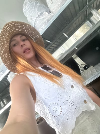 A young woman with long, reddish-blonde hair and fair skin wears a white, eyelet-lace top and a straw hat. She is looking at the camera with her tongue sticking out, appearing playful. A dark-colored pair of sunglasses is tucked into the front of her top. The image is a selfie taken from a low angle, giving a sense of looking up at her.
The background suggests an indoor setting, possibly under construction or in a warehouse. Metal framing, commonly used in ceiling construction, is visible. There are also large, white plastic-covered objects, which could be furniture or materials covered for protection. The lighting appears to be bright and somewhat diffused, possibly from overhead artificial lights. The overall impression is a candid and informal moment captured by the subject herself.