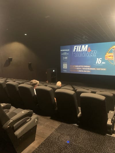 The image shows an interior view of a cinema auditorium in Sinalunga, Italy, during an intermission. The screen displays an advertisement for "Film & Dinner," indicating a movie screening combined with a meal. The advertisement features the price of 16.90€ for a cinema ticket that includes a pizza, smashburger, hot dog, and a 0.5L drink. A timer on the screen shows "1:38" for the remaining intermission time. Several rows of empty, dark-colored cinema seats are visible in the foreground and mid-ground. Two individuals are seated in the mid-ground, facing away from the camera, towards the screen. The environment appears dim, typical of a cinema setting. The floor in the foreground is carpeted with a dark pattern of white dots, and in the background, it appears to be wooden flooring.