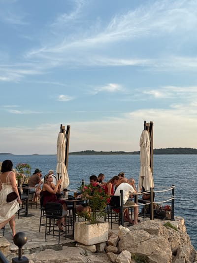 A group of people are dining at tables set up on a rocky terrace overlooking the Adriatic Sea in Primošten, Croatia. The sky is a pale blue with wispy clouds, and the sun appears to be setting, casting a warm glow on the scene. The terrace is paved with stone and features several outdoor tables and chairs. Two large, cream-colored umbrellas are partially furled, providing some shade.

Several individuals are seated at the tables, engaged in conversation and enjoying their meals. One woman, wearing a white dress, is standing and walking away from the camera, carrying a dark bag. Another woman, in a maroon dress, is seated at a table, looking at her phone. A man in a white t-shirt and shorts is also seated, resting his head on his hand. Other diners are visible further back, some in casual attire.

The sea is a deep blue, with gentle waves lapping against the rocky shore. In the distance, small islands or peninsulas are visible, covered in lush green vegetation. The atmosphere appears relaxed and casual, suggesting a pleasant evening meal in a scenic location. The overall impression is one of a charming seaside restaurant on a beautiful evening.