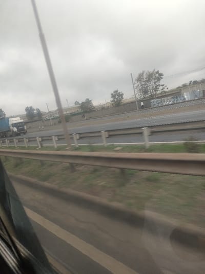 The image displays an overcast daytime scene of a multi-lane highway in Gatongora ward, Kenya, captured from a moving vehicle. A brown metal guardrail occupies the mid-foreground, with patches of green grass between it and the lower edge of the frame, which shows a blurred dark window section. The highway features at least three visible asphalt lanes, separated by concrete barriers. In the leftmost lane, a white Scania semi-truck with a blue trailer is observed moving in the same direction as the camera. Beyond the highway, a substantial concrete retaining wall runs horizontally, topped with some fencing. Behind this wall, a series of industrial or commercial buildings are visible, interspersed with trees. Electrical utility poles and overhead power lines traverse the background. Two indistinct human figures are present near the concrete wall; one figure, wearing a white top, stands near a utility pole. Further to the right, large cylindrical storage tanks are partially visible behind the wall.