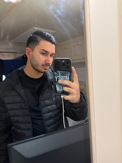 An individual is depicted in a mirror selfie taken indoors in Fowlis Easter, United Kingdom. The subject is a male with dark hair styled with a fade, a trimmed beard, and a mustache. He is wearing a black long-sleeved shirt under a black puffer jacket featuring "BOSS" on the left chest area. He holds a dark-colored smartphone with his right hand, positioned in front of his torso, capturing his reflection. The phone case displays the phrase "TIME IS MONEY" and a stack of hundred-dollar bills, reversed due to the mirror reflection. A white charging cable is connected to the bottom of the phone.
The reflected environment behind the subject includes light-colored kitchen-style cabinets in the upper right, and a ceiling with a rectangular vent or fixture in the upper left. Further left in the background reflection, dark fabric or clothing hangs. A dark, flat, rectangular object, possibly a monitor or desk surface, is visible in the lower foreground of the reflection. The wall immediately adjacent to the mirror on the right is a plain light color.