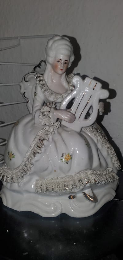 A white porcelain figurine of a woman with a powdered wig and an ornate dress sits holding a lyre. The woman's eyes are looking forward, and her lips are painted red. The dress has ruffled trim with gold accents. Small yellow flowers are painted on the dress. The base of the figurine is white and wavy, with gold accents on the edge. The figurine is on a dark, reflective surface. The background is a light-colored wall with some shadows.
